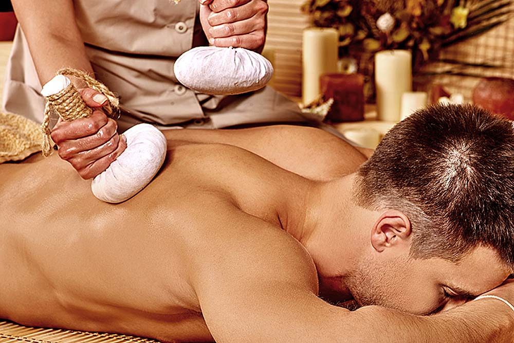 Hot Stone Massage Ein Mann liegt entspannt auf dem Bauch, während eine Therapeutin mit Kräuterstempeln eine Massage auf seinem Rücken durchführt.